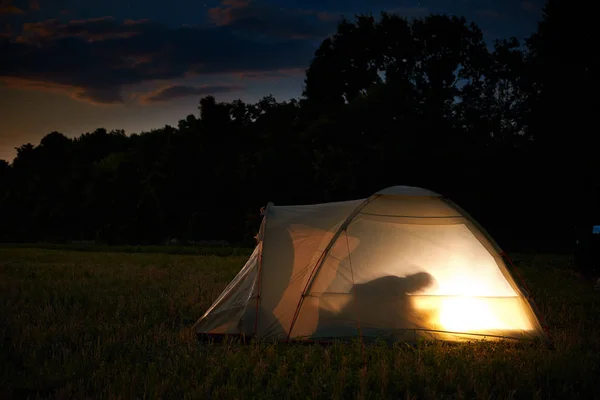 Traveling and camping concept - camp tent at night under a sky full of stars. Orange illuminated tent with a person inside. Beautiful nature - field, forest, plain. Moon and moonlight — Stock Photo, Image