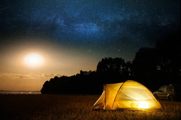 Concepto de viaje y camping - tienda de campaña en la noche bajo un cielo lleno de estrellas. Tienda iluminada naranja y coche. Hermosa naturaleza - campo, bosque, llanura. Luna y luz de luna —  Fotos de Stock