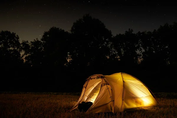 Traveling and camping concept - camp tent at night under a sky full of stars. Orange illuminated tent. Beautiful nature - field, forest, plain. Moon and moonlight