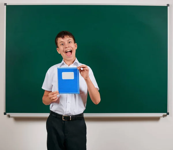 Portrait of a school boy posing with blue exercise book on blackboard background - back to school and education concept — Stock Photo, Image