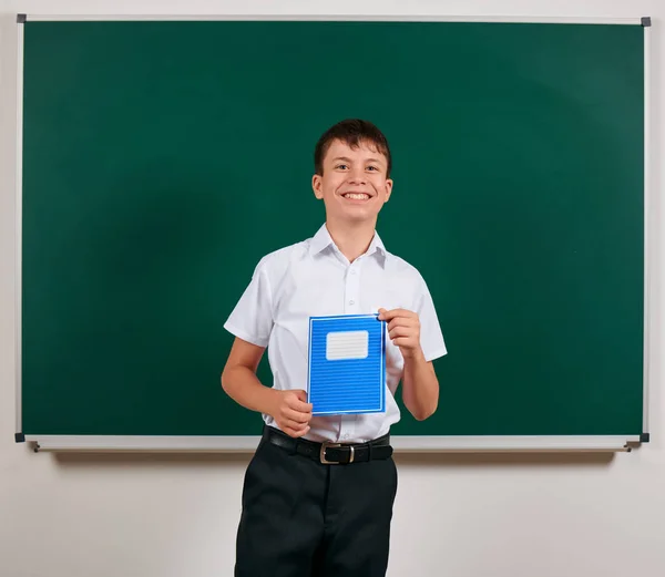 Портрет школьника, позирующего с синей тетрадью на фоне доски - возвращение к школьной и образовательной концепции — стоковое фото