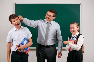 Bir öğretmenin portresi kötü disiplinve mutlu bir kız öğrenci olan okul çocuğunun kulağını kapmak. Kara tahta arka plan üzerinde egzersiz kitapları, kalemler, kalemler ve diğer okul malzemeleri ile poz