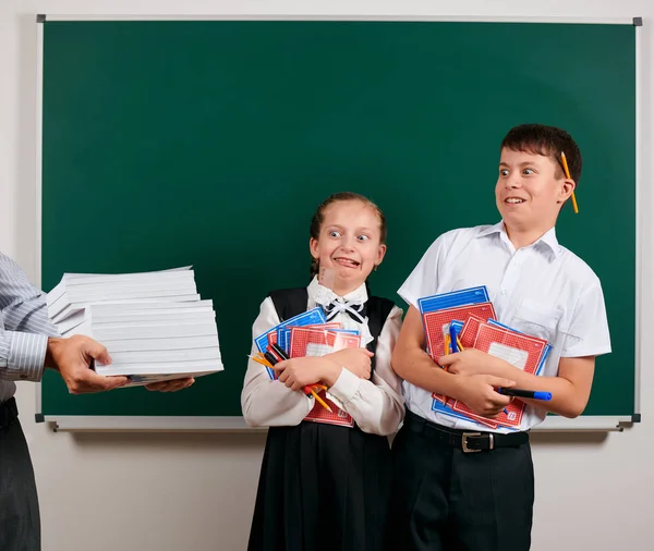 Emotional portrait of a schoolboy and schoolgirl get a lot of books, posing with notebooks, pens, pencils and other school supplies on blackboard background - back to school and education concept — Stock Photo, Image