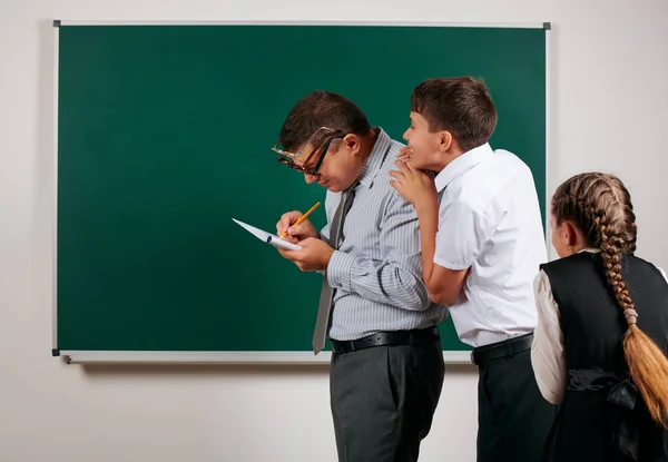 Portrait of a teacher checking homework, reading school exercise books, schoolboy and schoolgirl with old fashioned eyeglasses posing on blackboard background - back to school and education concept — Stock Photo, Image