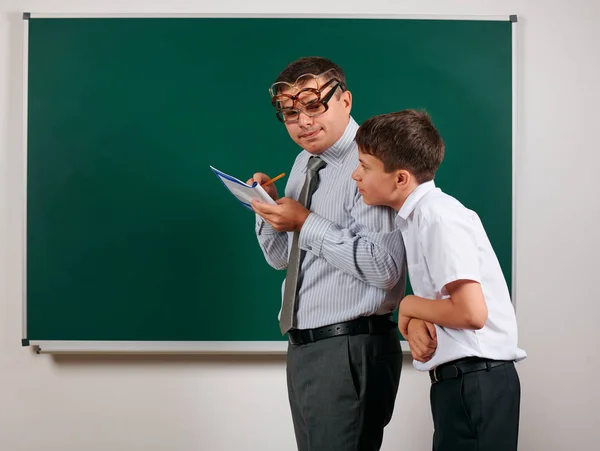 Portrait of a teacher checking homework, reading school exercise books and schoolboy, posing on blackboard background - back to school and education concept — Stock Photo, Image
