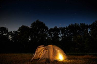 Seyahat ve kamp konsepti - yıldız dolu bir gökyüzü altında geceleri kamp çadırı. Turuncu ışıklı çadır. Güzel doğa - alan, orman, düz. Ay ve ay ışığı