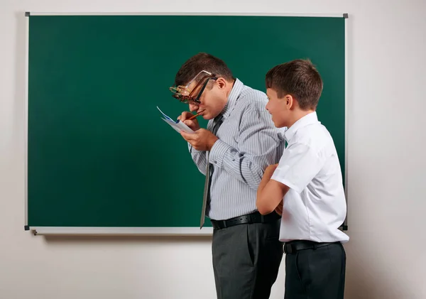 Portrait of a teacher checking homework, reading school exercise books and schoolboy posing on blackboard background - back to school and education concept — Stock Photo, Image