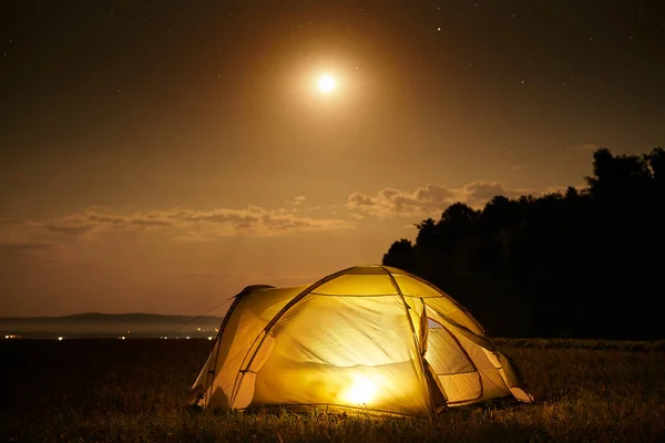 Traveling and camping concept - camp tent at night under a sky full of stars. Orange illuminated tent. Beautiful nature - field, forest, plain. Moon and moonlight