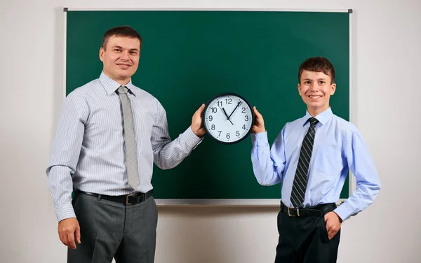 Ritratto di un uomo e di un ragazzo vestiti in abiti da lavoro vicino alla lavagna sfondo, mostrano orologio - apprendimento e concetto di istruzione — Foto Stock