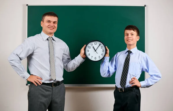Portrait of a man and boy dressed in a business suits near blackboard background, they show clock - learning and education concept