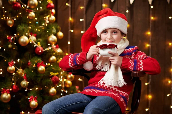 Menina ajudante alegre do Papai Noel com xícara de chá sentado no interior perto da árvore de xmas decorada com luzes, vestido com suéter vermelho - Feliz Natal e Boas Festas ! — Fotografia de Stock