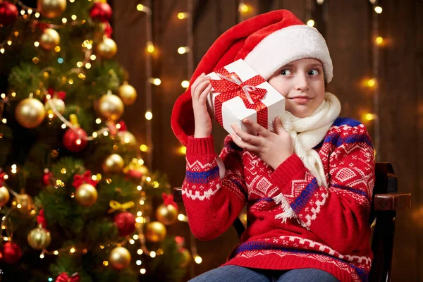 Fröhliches Weihnachtsmann-Helfermädchen mit Geschenkbox sitzt drinnen neben geschmücktem Weihnachtsbaum mit Lichtern, gekleidet in rotem Pullover - frohe Weihnachten und frohe Feiertage! — Stockfoto