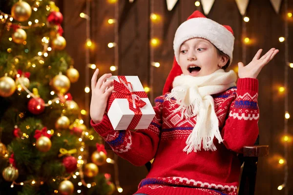 Menina ajudante alegre do Papai Noel com caixa de presente sentada no interior perto da árvore de xmas decorada com luzes, vestida com suéter vermelho - Feliz Natal e Boas Festas ! — Fotografia de Stock