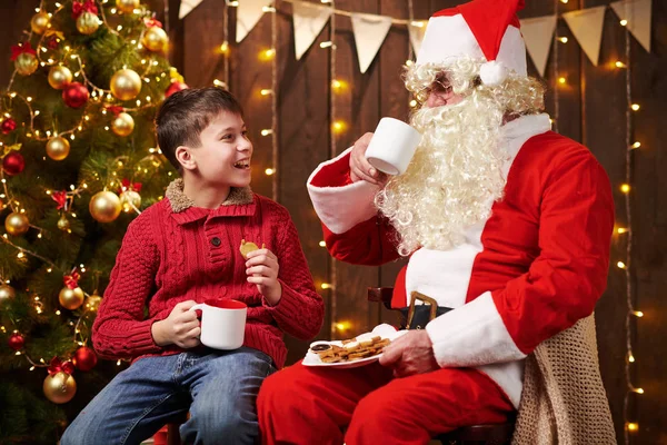Санта Клаус і хлопчик п "ють чай, їдять печиво, розмовляють і сидять у приміщенні біля прикрашеного ксами дерева з вогнями - Веселого Різдва і Веселих свят.! — стокове фото