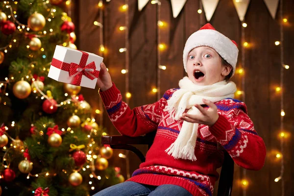 Joyeux santa helper girl avec boîte cadeau assis à l'intérieur près décoré arbre de Noël avec des lumières, vêtu de pull rouge Joyeux Noël et Joyeuses Fêtes ! — Photo