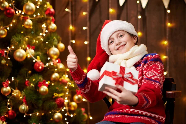 Menina ajudante alegre do Papai Noel com caixa de presente sentada no interior perto da árvore de xmas decorada com luzes, vestida com suéter vermelho - Feliz Natal e Boas Festas ! — Fotografia de Stock