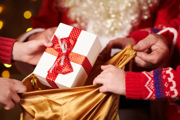 Babbo Natale apre una borsa e fa regali ai bambini, seduti vicino all'albero di Natale decorato con luci Buon Natale e Buone Feste ! — Foto Stock