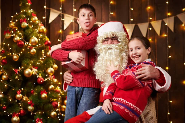 Санта Клаус і хлопчик і дівчинка, що тиняються в приміщенні біля прикрашеного дерева ксам зі світлом, вони розмовляють, усміхаються і приймають подарунки - Веселого Різдва і Веселих свят.! — стокове фото