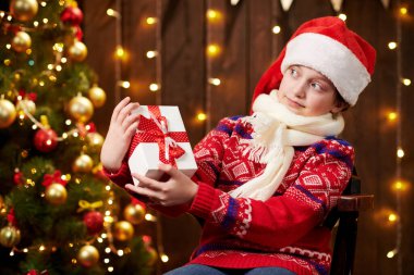 Cheerful santa helper girl with gift box sitting indoor near decorated xmas tree with lights, dressed in red sweater - Merry Christmas and Happy Holidays!