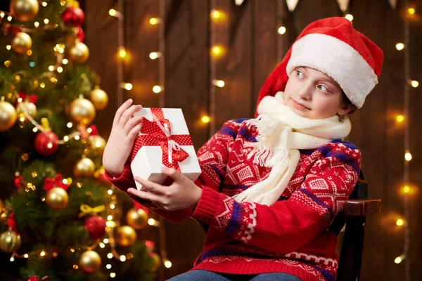 Cheerful santa helper girl with gift box sitting indoor near decorated xmas tree with lights, dressed in red sweater - Merry Christmas and Happy Holidays! — стокове фото