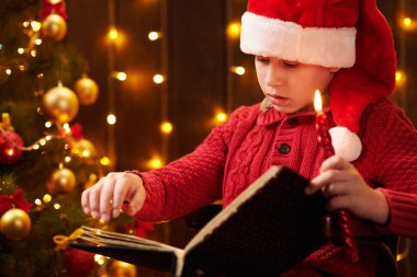 Genç çocuk kitap okuyor, kapalı bir yerde Noel Baba yardımcısı gibi giyinmiş, ışıklarla süslenmiş Xmas ağacının yanında oturuyor.!