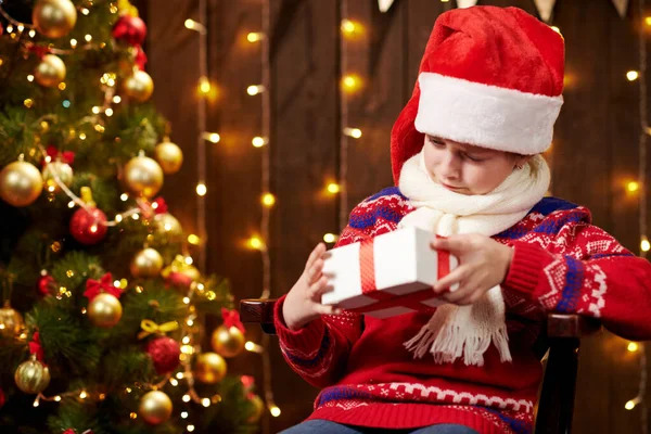 Allegro Babbo Natale aiutante ragazza con scatola regalo seduto al coperto vicino all'albero di Natale decorato con luci, vestito con maglione rosso Buon Natale e Buone Feste ! — Foto Stock