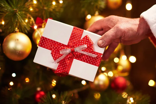 Closeup of Santa Claus hands with gift box, sitting indoor near decorated xmas tree with lights - Merry Christmas and Happy Holidays! — Stock Photo, Image