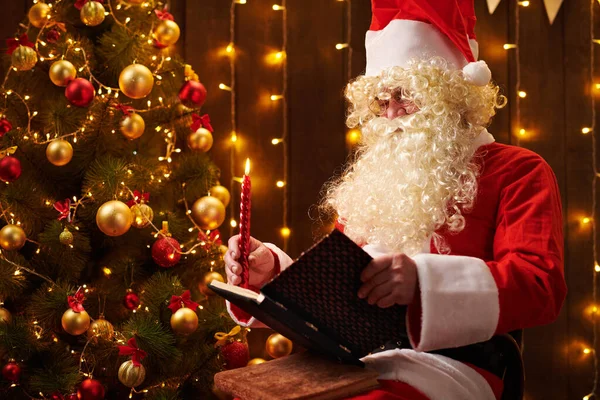 Libro de lectura de Papá Noel, sentado en el interior cerca del árbol de Navidad decorado con luces - Feliz Navidad y Felices Fiestas ! — Foto de Stock