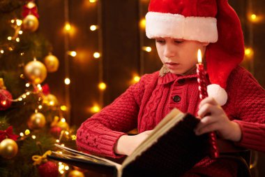 Genç çocuk kitap okuyor, kapalı bir yerde Noel Baba yardımcısı gibi giyinmiş, ışıklarla süslenmiş Xmas ağacının yanında oturuyor.!