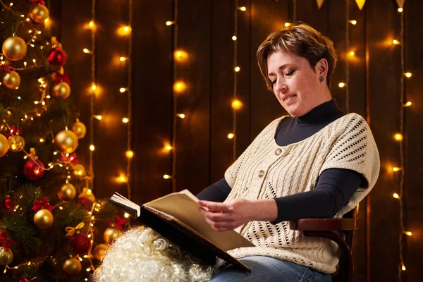 Krásná žena čtení knihy, sedí uvnitř v blízkosti zdobené vánoční strom se světly - Veselé Vánoce a šťastné svátky! — Stock fotografie
