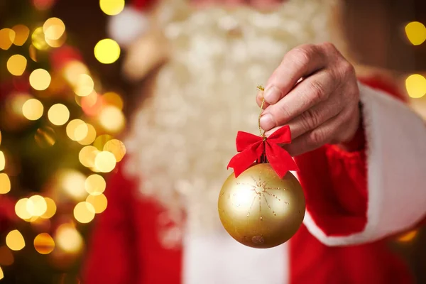 Sluiten van de kerstman handen met kerstbal decoratie, zittend binnen in de buurt van versierde kerstboom met verlichting - Vrolijk kerstfeest en fijne feestdagen! — Stockfoto