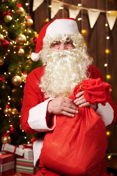 Père Noël posant avec un sac de cadeaux, assis à l'intérieur près décoré arbre de Noël avec des lumières Joyeux Noël et Joyeuses Fêtes ! — Photo