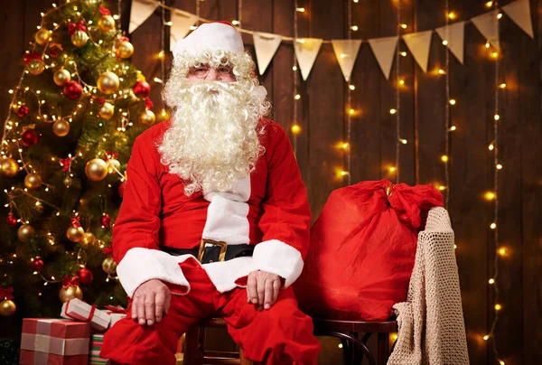 Père Noël posant avec un sac de cadeaux, assis à l'intérieur près décoré arbre de Noël avec des lumières Joyeux Noël et Joyeuses Fêtes ! — Photo