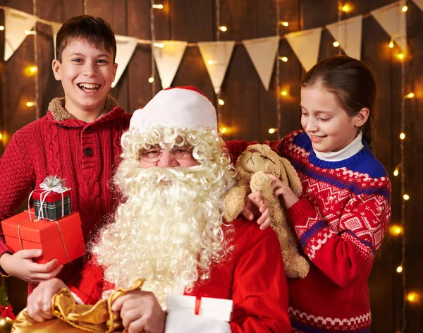 Санта Клаус відкриває сумку і дарує дітям подарунки, сидячи в приміщенні біля прикрашеного ксами дерева з вогнями - Веселого Різдва і Веселих свят.! — стокове фото