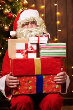 Noel Baba bir sürü hediyeyle poz veriyor, kapalı bir yerde süslü Noel ağacının yanında oturuyor - Mutlu Noeller ve mutlu tatiller!