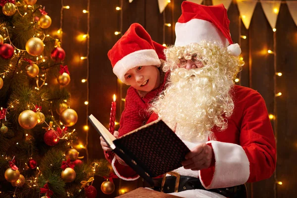 Père Noël et santa aide garçon livre de lecture, assis à l'intérieur près décoré arbre de Noël avec des lumières Joyeux Noël et Joyeuses Fêtes ! — Photo