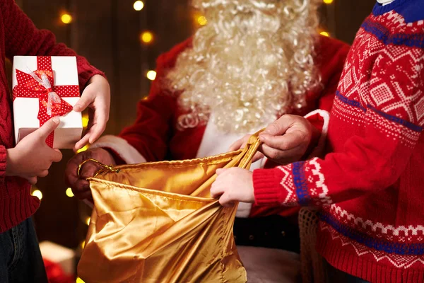 サンタクロースは袋を開き、子供たちに贈り物を与え、装飾されたクリスマスツリーの近くにライトで屋内に座っています-メリークリスマスとハッピーホリデー! — ストック写真