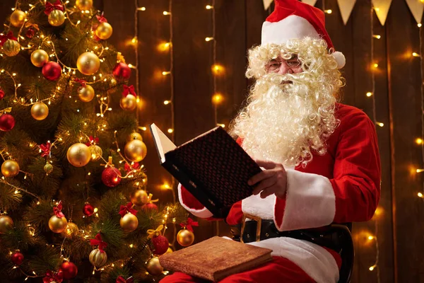 サンタクロース読書本、照明付きの装飾されたクリスマスツリーの近くに屋内に座って-メリークリスマスとハッピーホリデー! — ストック写真