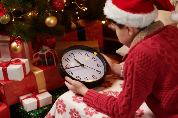 Adolescent garçon attendant le Père Noël et regarder l'horloge, couché à l'intérieur près décoré arbre de Noël avec des lumières, habillé comme aide Père Noël Joyeux Noël et Joyeuses Fêtes ! — Photo