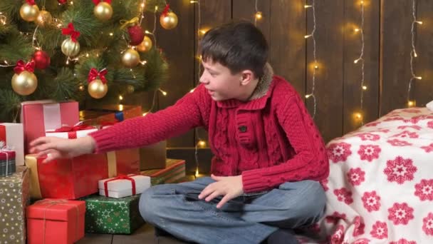 男孩坐在装饰过的圣诞树旁 点着灯 准备礼物 圣诞快乐和节日快乐 — 图库视频影像