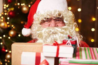 Noel Baba bir sürü hediyeyle poz veriyor, kapalı bir yerde süslü Noel ağacının yanında oturuyor - Mutlu Noeller ve mutlu tatiller!
