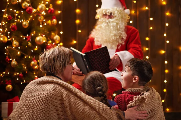 Санта Клаус читає книжку для сім'ї. Мати з дітьми сидять у приміщенні біля прикрашеного ксам дерева з сяйвом - Веселого Різдва і Веселих свят.! — стокове фото