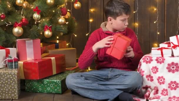 男の子はライトとギフトを得る装飾されたクリスマスツリーの近くに屋内に座っています メリークリスマスとハッピーホリデー — ストック動画