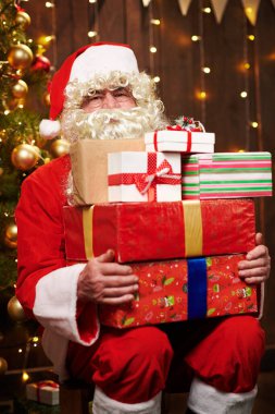 Noel Baba bir sürü hediyeyle poz veriyor, kapalı bir yerde süslü Noel ağacının yanında oturuyor - Mutlu Noeller ve mutlu tatiller!