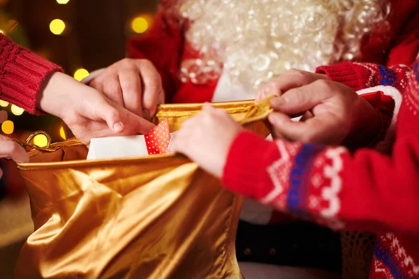 Père Noël ouvre un sac et donne des cadeaux aux enfants, assis à l'intérieur près de l'arbre de Noël décoré avec des lumières Joyeux Noël et Joyeuses Fêtes ! — Photo