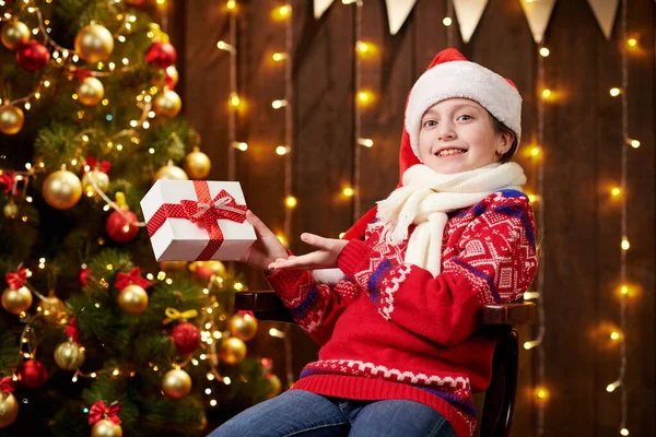 Cheerful santa helper girl with gift box sitting indoor near decorated xmas tree with lights, dressed in red sweater - Merry Christmas and Happy Holidays! — Stock fotografie