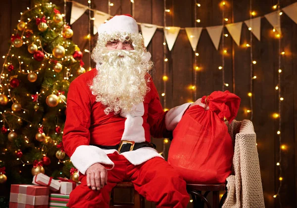 Babbo Natale in posa con sacchetto di regali, seduto vicino all'albero di Natale decorato con luci - Buon Natale e Buone Feste ! — Foto Stock