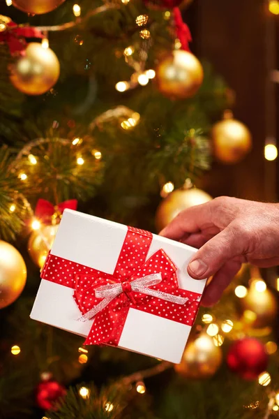 Close-up van de kerstman handen met geschenkdoos, zittend binnen in de buurt van versierde kerstboom met verlichting - Vrolijk kerstfeest en fijne feestdagen! — Stockfoto