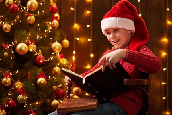 Tiener jongen leesboek, zittend binnen in de buurt van versierde kerstboom met verlichting, verkleed als Santa helper - Vrolijk kerstfeest en fijne feestdagen! — Stockfoto
