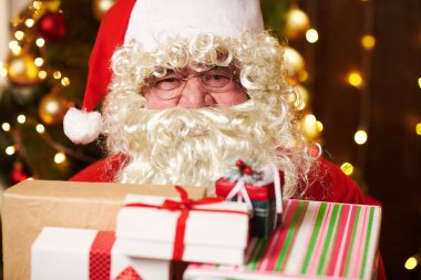 Noel Baba bir sürü hediyeyle poz veriyor, kapalı bir yerde süslü Noel ağacının yanında oturuyor - Mutlu Noeller ve mutlu tatiller!